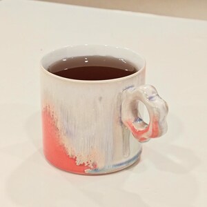 May include: A ceramic mug with a unique floral-shaped handle. The mug is white with a red and blue color wash on the exterior and filled with a dark liquid. The mug is designed for hot beverages.