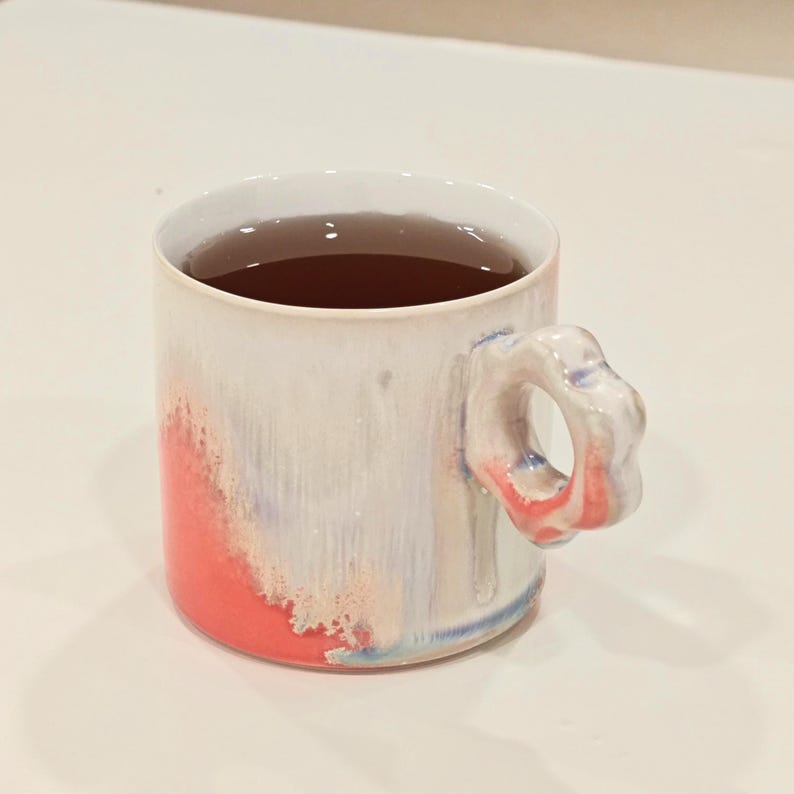 May include: A ceramic mug with a unique floral-shaped handle. The mug is white with a red and blue color wash on the exterior and filled with a dark liquid. The mug is designed for hot beverages.