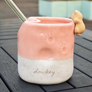May include: A pink and white ceramic mug with paw prints and a small bear figurine attached. The mug has the word "Lowkey" written on it and includes a metal straw.