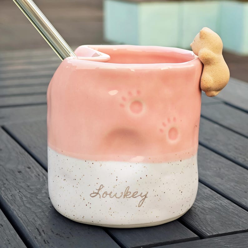May include: A pink and white ceramic mug with paw prints and a small bear figurine attached. The mug has the word "Lowkey" written on it and includes a metal straw.
