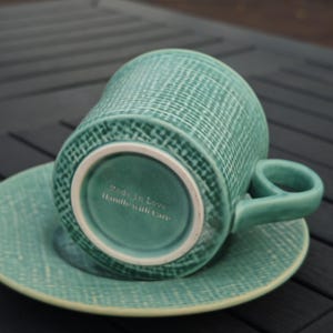 May include: A teal ceramic teacup and saucer set. The cup is upside down, revealing the base with the text "Made in Love Handle with Care". The set has a textured woven pattern. The handle is a rounded shape.