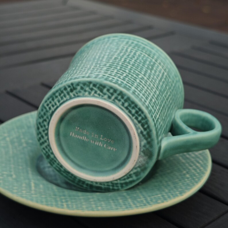 May include: A teal ceramic teacup and saucer set. The cup is upside down, revealing the base with the text "Made in Love Handle with Care". The set has a textured woven pattern. The handle is a rounded shape.
