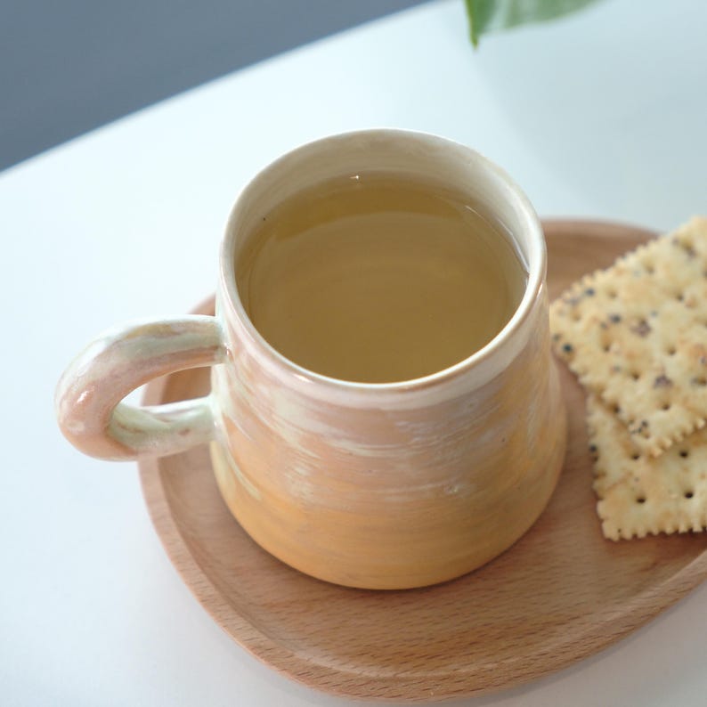 May include: A handmade ceramic mug with a light orange and cream glaze, filled with tea. The mug sits on a wooden tray with crackers. The mug has a rounded handle and a wide mouth. The tea is a light amber color.