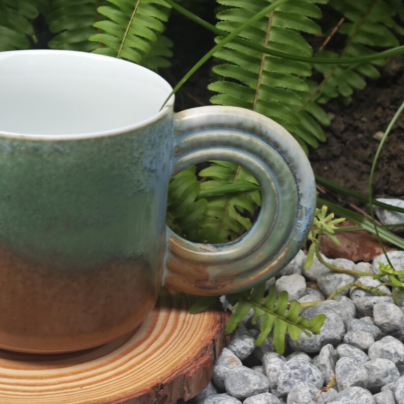 K&ouml;nnte beinhalten: Eine Keramik-Tasse mit einem einzigartigen Griffdesign. Die Tasse hat einen Farbverlauf, der von Gr&uuml;n nach Braun &uuml;bergeht. Der Griff besteht aus einer Reihe konzentrischer Kreise in Blau- und Gr&uuml;nt&ouml;nen. Die Tasse steht auf einem Holzuntersetzer.