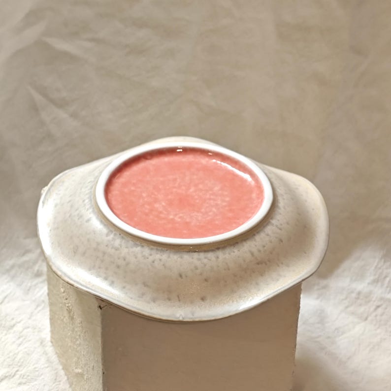 May include: A ceramic dish with a light beige exterior and a coral-colored interior. The dish has a unique, irregular shape with a slightly raised rim. The interior is a solid coral color, while the exterior has a speckled texture.