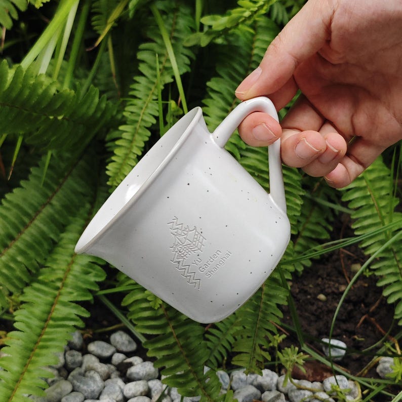 May include: A white speckled ceramic mug with a curved handle. The mug has an embossed design and the text "Yu Garden Shanghai". The mug is held in a hand against a background of green foliage and small rocks.
