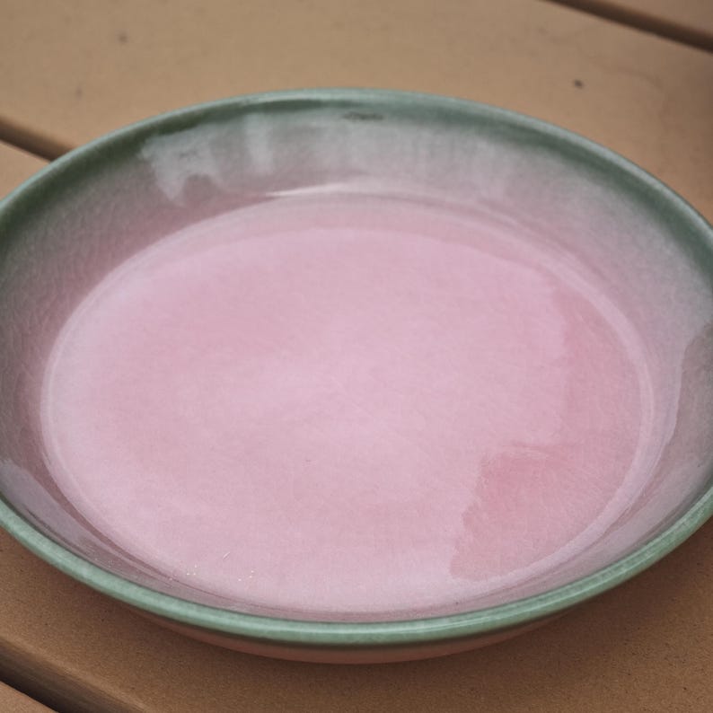 Puede incluir: Un plato redondo de cer&aacute;mica que presenta un centro de color rosa claro brillante y suave que transiciona hacia un borde de color verde salvia apagado. La superficie tiene un acabado esmaltado reflectante con variaciones.