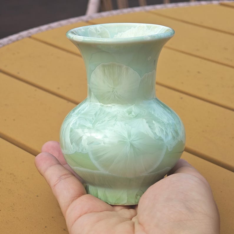 Puede incluir: Un peque&ntilde;o jarr&oacute;n de cer&aacute;mica verde claro con un cuerpo bulboso y un borde ensanchado. El jarr&oacute;n tiene un patr&oacute;n cristalino &uacute;nico en blanco, creando una apariencia texturizada. El jarr&oacute;n se sostiene en una mano, mostrando su tama&ntilde;o y forma.