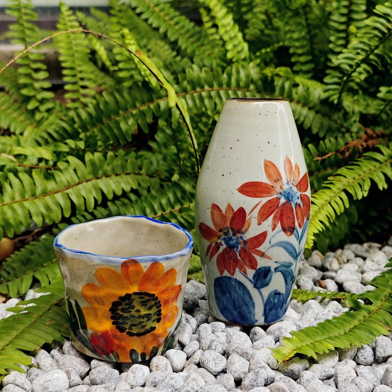 May include: Two ceramic vases with floral designs. One is a small cup with a sunflower, the other is a tall vase with red and blue flowers. The vases are on a bed of white stones, with green foliage in the background.