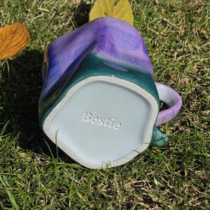 May include: A handmade ceramic mug with a unique shape and a colorful glaze. The mug features a blend of purple, teal, and green hues. The word "Bestie" is embossed on the bottom of the mug. The mug is sitting on a bed of green grass.