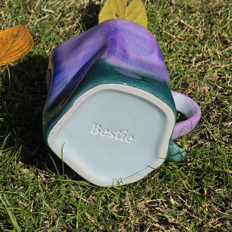 May include: A handmade ceramic mug with a unique shape and a colorful glaze. The mug features a blend of purple, teal, and green hues. The word "Bestie" is embossed on the bottom of the mug. The mug is sitting on a bed of green grass.