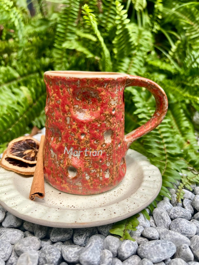 May include: A handmade ceramic mug with a textured red and brown surface, the word "Martian" is written on the side. The mug sits on a speckled white plate with a cinnamon stick and dried orange slice.
