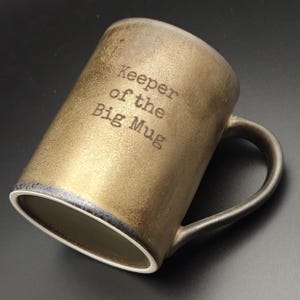 Puede incluir: Una taza cilíndrica de cerámica con un acabado metálico en oro y bronce. El exterior presenta el texto impreso en negro "Keeper of the Big Mug" en una fuente estilo máquina de escribir. La taza tiene un asa oscura y un borde interior blanco.