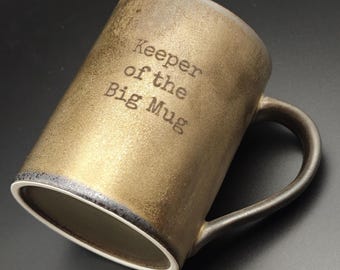 May include: A cylindrical ceramic mug with a metallic gold and bronze finish. The exterior features the black printed text "Keeper of the Big Mug" in a typewriter-style font. The mug has a dark handle and a white interior rim.