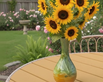May include: A bouquet of sunflowers in a green and yellow vase. The sunflowers have bright yellow petals and dark brown centers. The vase is on a round, light brown table. The background shows a garden with a fence and rose bushes.