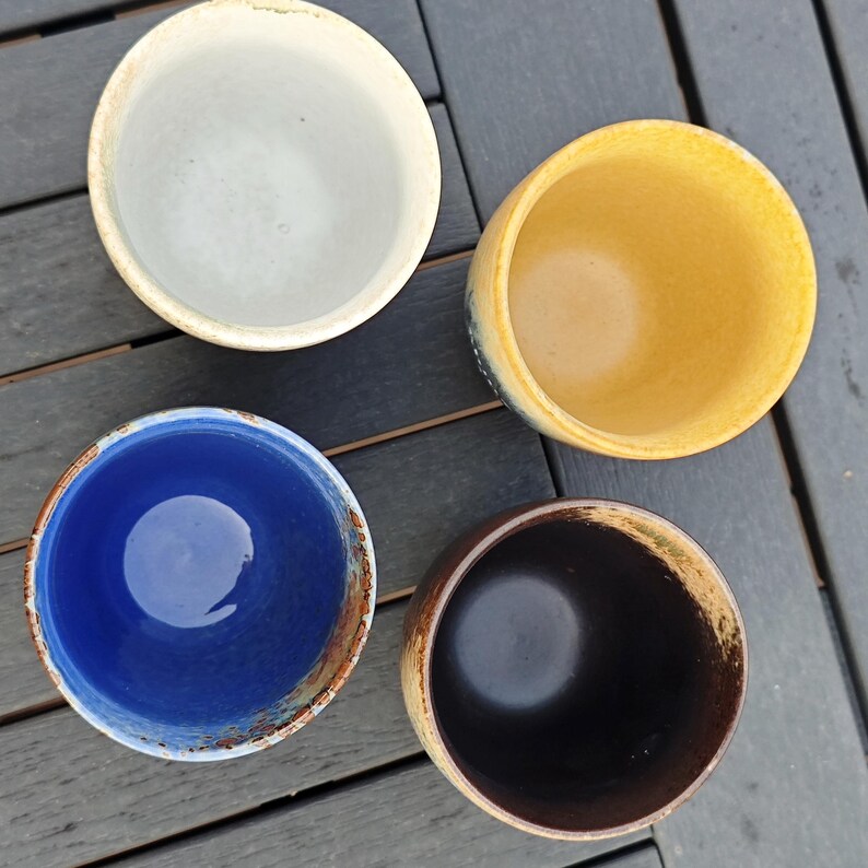 Puede incluir: Vista superior de cuatro cuencos de cer&aacute;mica. Los cuencos son de diferentes colores: blanco roto, amarillo, azul y marr&oacute;n. Tienen un aspecto r&uacute;stico y artesanal, con textura visible y variaciones de color. Los cuencos est&aacute;n colocados sobre una superficie de madera oscura.