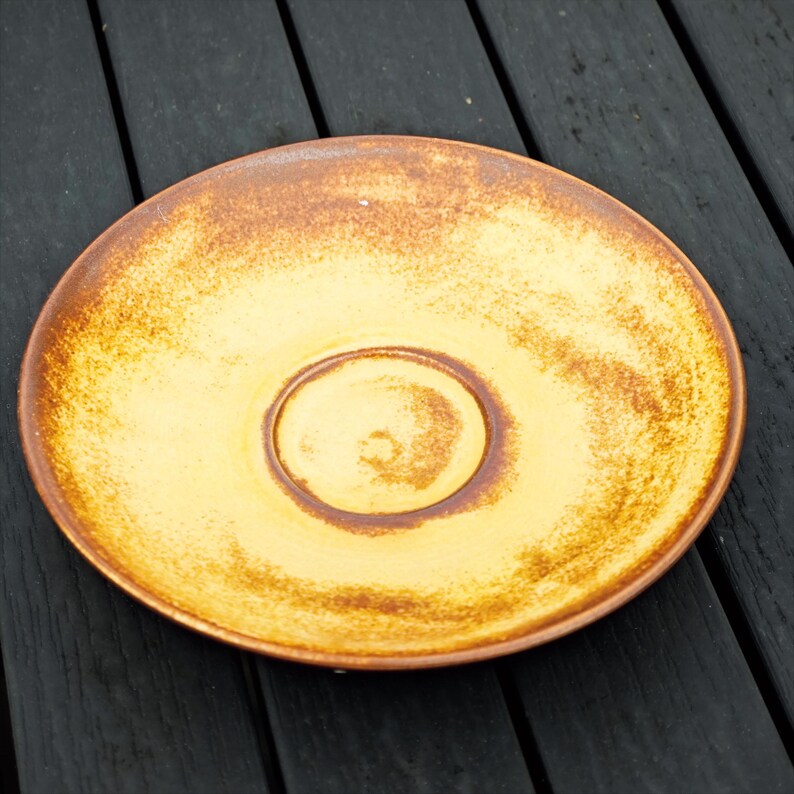 May include: A ceramic saucer with a warm, earthy color palette. The center is a light yellow, encircled by a darker brown ring. The rim and outer edges are a rich, mottled brown, creating a rustic, handcrafted aesthetic. The saucer is designed for a cup.