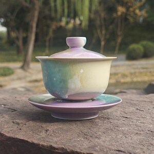 May include: A ceramic tea set with a pastel color scheme. The set includes a lidded gaiwan tea bowl and a matching saucer. The bowl and saucer have a gradient of colors, including blue, green, yellow, and purple. The tea set is placed on a stone surface.