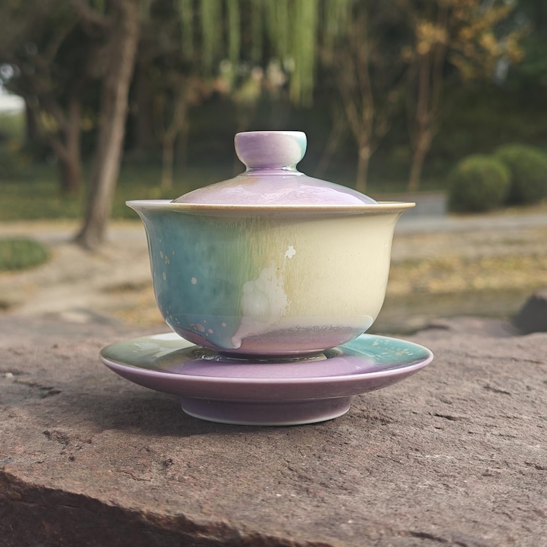 May include: A ceramic tea set with a pastel color scheme. The set includes a lidded gaiwan tea bowl and a matching saucer. The bowl and saucer have a gradient of colors, including blue, green, yellow, and purple. The tea set is placed on a stone surface.