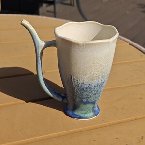 May include: A handmade ceramic mug with a unique handle design. The mug features a white and blue glaze, with a textured appearance. The handle is a curved shape, and the mug has a flared rim. The mug is suitable for hot or cold beverages.
