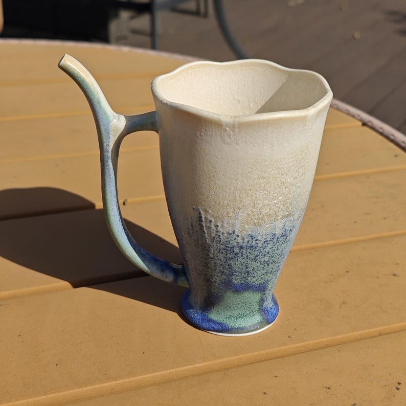 May include: A handmade ceramic mug with a unique handle design. The mug features a white and blue glaze, with a textured appearance. The handle is a curved shape, and the mug has a flared rim. The mug is suitable for hot or cold beverages.