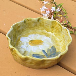 May include: A hand-painted ceramic bowl with a daisy design. The bowl has a scalloped edge and a yellow-green exterior. The daisy has white petals and a yellow center, with blue leaves. A sprig of white flowers is in the background.