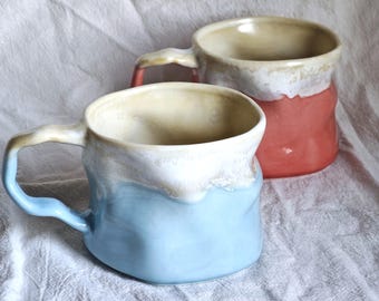 May include: Two handmade ceramic mugs. One is light blue with a white rim and handle, the other is red with a white rim and handle. Both mugs have a textured, organic shape and a beige interior.