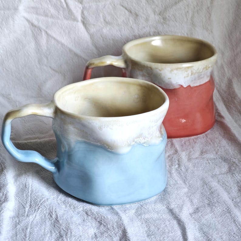 May include: Two handmade ceramic mugs. One is light blue with a white rim and handle, the other is red with a white rim and handle. Both mugs have a textured, organic shape and a beige interior.