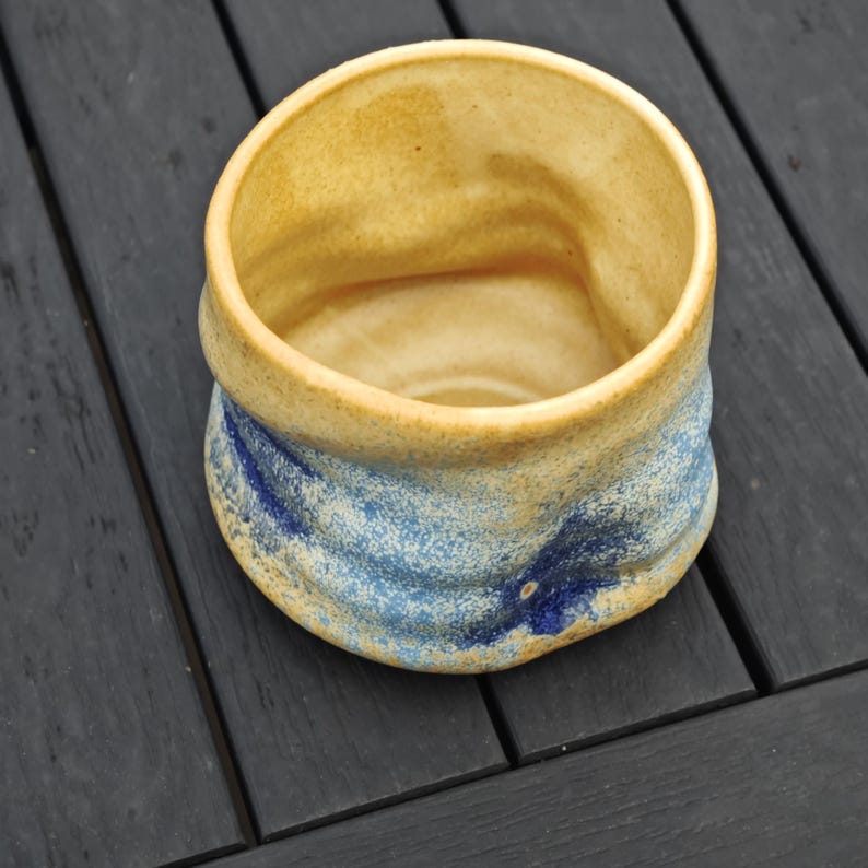 Peut inclure: Tasse en c&eacute;ramique faite &agrave; la main avec un design unique. L'int&eacute;rieur et le bord sont de couleur beige clair, tandis que l'ext&eacute;rieur pr&eacute;sente des tons bleus et beiges. La forme l&eacute;g&egrave;rement irr&eacute;guli&egrave;re de la tasse ajoute &agrave; son charme artisanal.