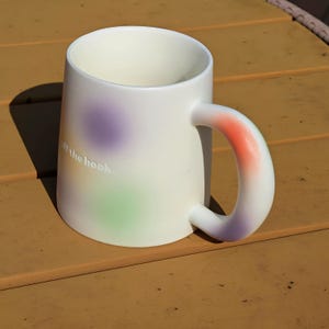 K&ouml;nnte beinhalten: Eine wei&szlig;e Keramik-Tasse mit einem abgerundeten Henkel. Die Tasse hat einen Farbverlauf aus Lila, Gr&uuml;n und Orange. Der Text "off the hook" ist auf der Seite der Tasse aufgedruckt.