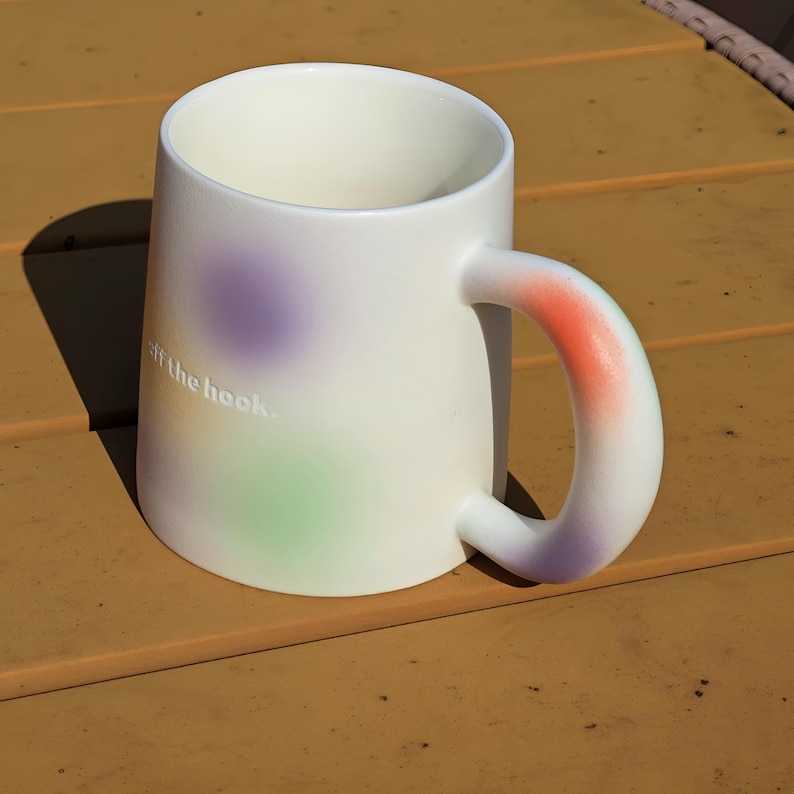 K&ouml;nnte beinhalten: Eine wei&szlig;e Keramik-Tasse mit einem abgerundeten Henkel. Die Tasse hat einen Farbverlauf aus Lila, Gr&uuml;n und Orange. Der Text "off the hook" ist auf der Seite der Tasse aufgedruckt.