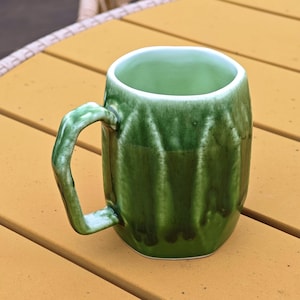 May include: A handmade green ceramic mug with a unique, textured design. The mug features a curved handle and a glossy finish, perfect for enjoying your favorite beverage. The interior is a lighter shade of green.