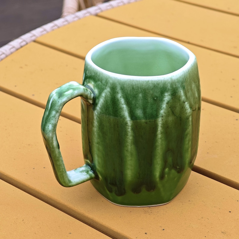 May include: A handmade green ceramic mug with a unique, textured design. The mug features a curved handle and a glossy finish, perfect for enjoying your favorite beverage. The interior is a lighter shade of green.