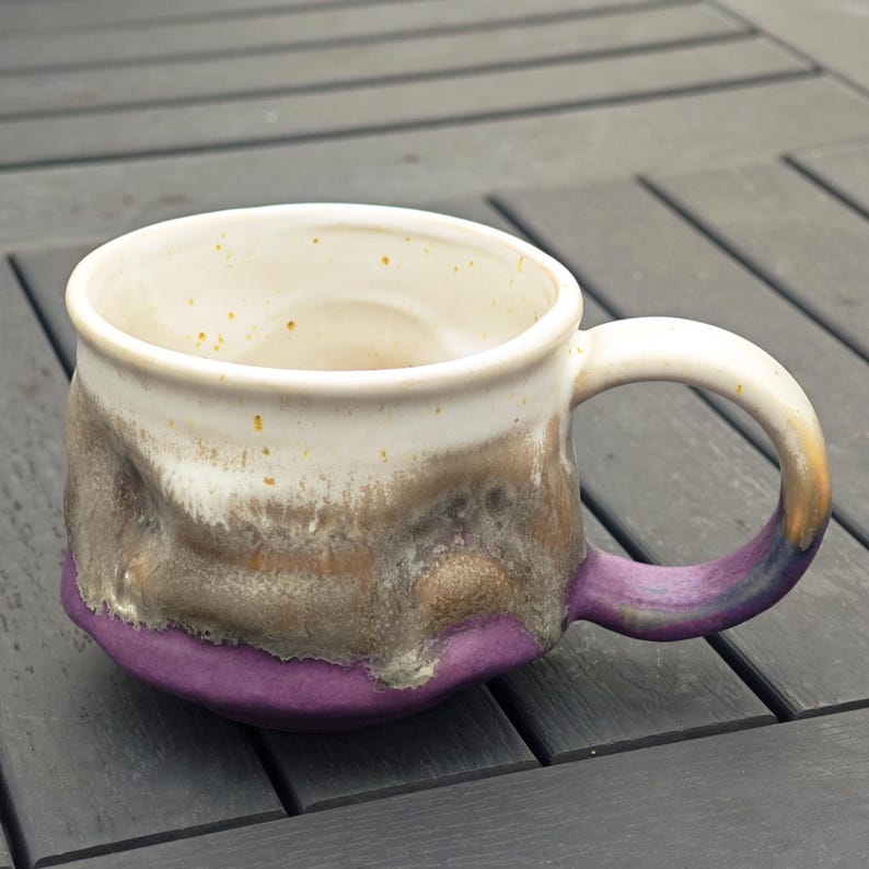 Op de afbeelding: Een handgemaakte keramische mok met een witte binnenkant en een getextureerde buitenkant. De mok heeft een kleurovergang, van wit naar bruin en paars. Het handvat is paars met een gouden accent. De mok is ongeveer 10 cm hoog.