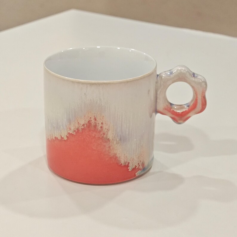May include: A ceramic mug with a unique flower-shaped handle. The mug features a gradient design, transitioning from a vibrant coral at the base to a white and light blue at the top. The interior is white.