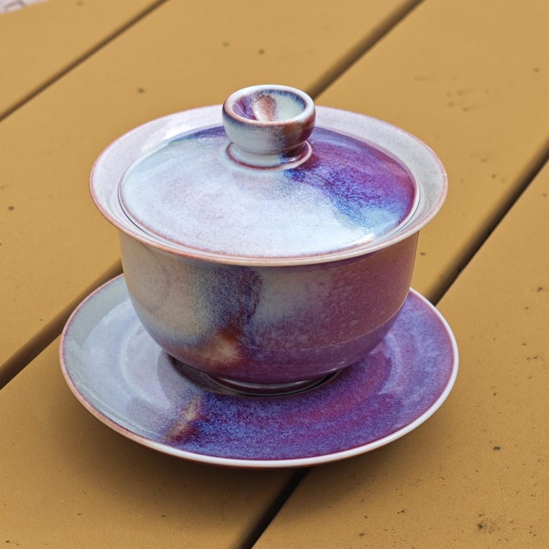 May include: A ceramic tea set with a purple, blue, and white glaze. The set includes a lidded bowl and a saucer. The lid has a small handle. The tea set is designed for serving tea.