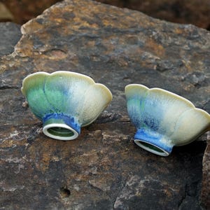 May include: Two ceramic bowls with a scalloped rim and a flared base. The bowls feature a blend of colors, including green, blue, and cream. The bowls are placed on a rough, dark stone surface.