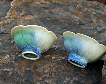 May include: Two ceramic bowls with a scalloped rim and a flared base. The bowls feature a blend of colors, including green, blue, and cream. The bowls are placed on a rough, dark stone surface.
