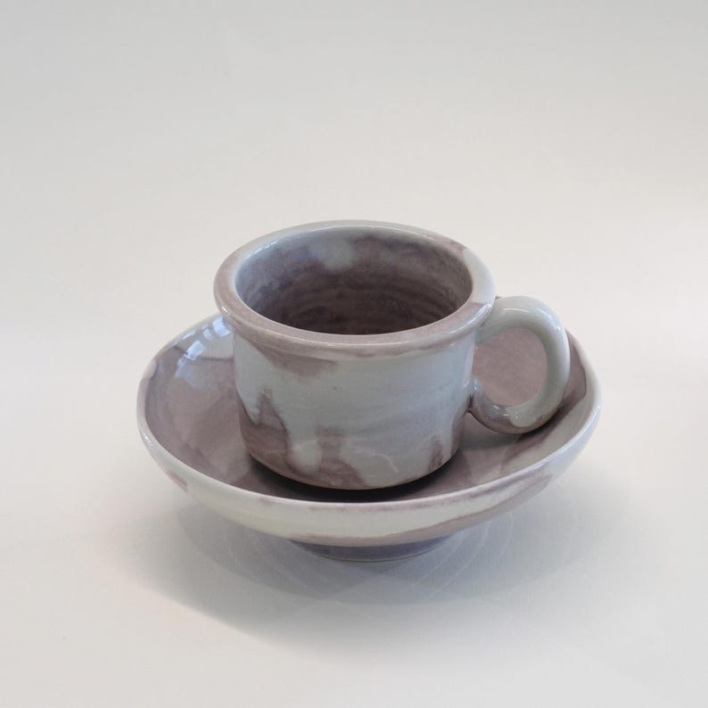 May include: A ceramic teacup and saucer set. The cup and saucer are a marbled blend of lavender and white. The cup has a small handle and sits on a matching saucer. The set is on a white surface.