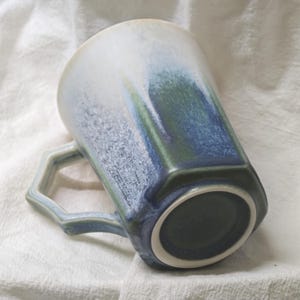May include: A ceramic mug with a unique geometric design. The mug features a gradient of colors, transitioning from white to blue and green. The handle has a distinctive shape, and the mug sits on a white surface.