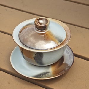 May include: A ceramic gaiwan tea set consisting of a bowl, a lid with a central knob, and a matching saucer. The pieces feature a light blue glaze with blended brown and black reactive glaze accents that create a gradient effect across the surfaces.