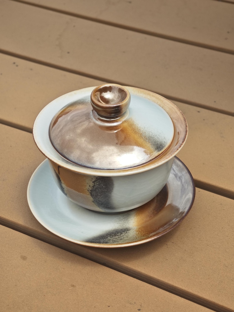 May include: A ceramic gaiwan tea set consisting of a bowl, a lid with a central knob, and a matching saucer. The pieces feature a light blue glaze with blended brown and black reactive glaze accents that create a gradient effect across the surfaces.