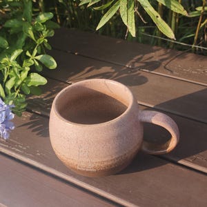 Może przedstawiać: Okrągły ceramiczny kubek z nakrapianą jasnor&oacute;żową g&oacute;rną częścią i ciemnobrązową podstawą. Na przedniej powierzchni kubka znajduje się mały, prosty rysunek liniowy przedstawiający kota, a po prawej stronie umieszczono zakrzywione ucho do trzymania.