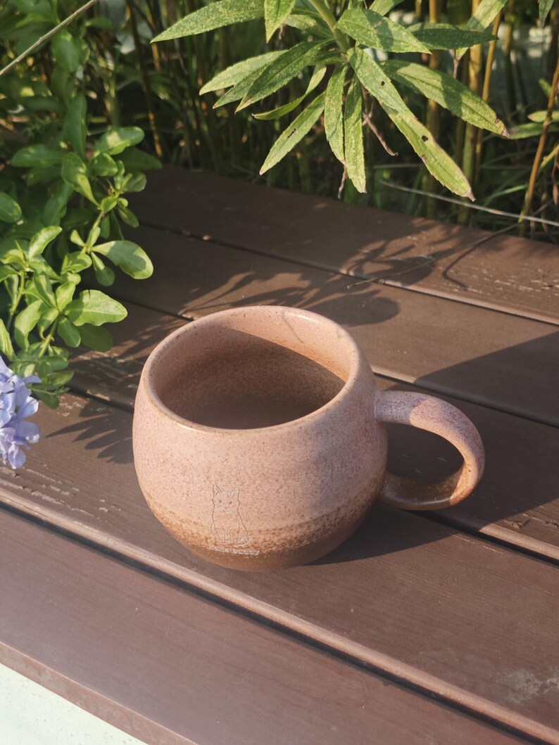 Może przedstawiać: Okrągły ceramiczny kubek z nakrapianą jasnor&oacute;żową g&oacute;rną częścią i ciemnobrązową podstawą. Na przedniej powierzchni kubka znajduje się mały, prosty rysunek liniowy przedstawiający kota, a po prawej stronie umieszczono zakrzywione ucho do trzymania.