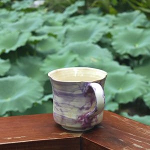 May include: A handmade ceramic mug with a white handle and a marbled design of purple and cream. The mug sits on a wooden surface with a blurred background of green lily pads.