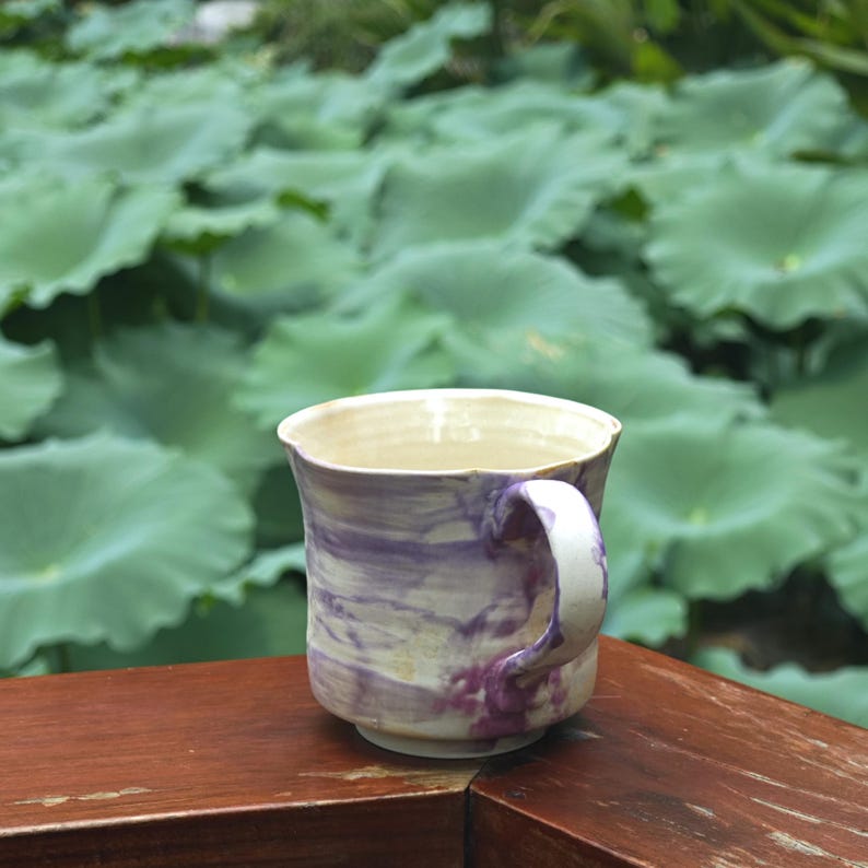 May include: A handmade ceramic mug with a white handle and a marbled design of purple and cream. The mug sits on a wooden surface with a blurred background of green lily pads.