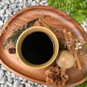 May include: A wooden serving tray holds a small, light-colored bowl filled with a dark liquid. Cinnamon sticks, a wooden ball, and a pine cone are arranged around the bowl. Ferns and small stones are in the background.