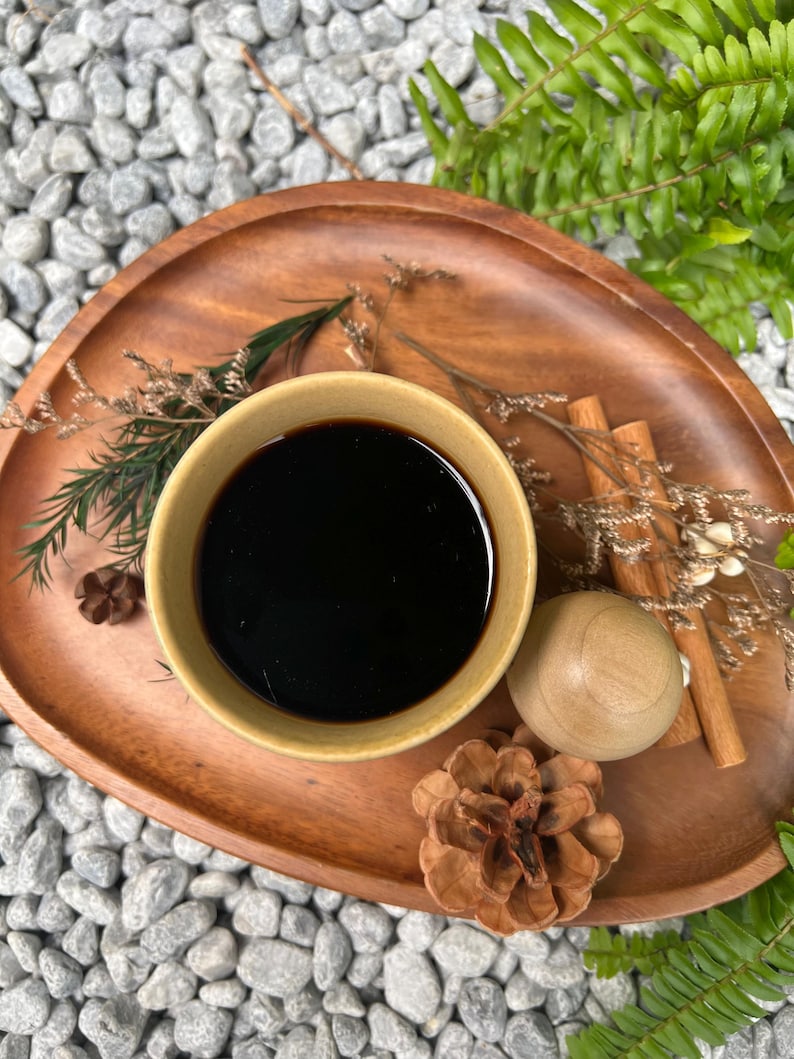 May include: A wooden serving tray holds a small, light-colored bowl filled with a dark liquid. Cinnamon sticks, a wooden ball, and a pine cone are arranged around the bowl. Ferns and small stones are in the background.