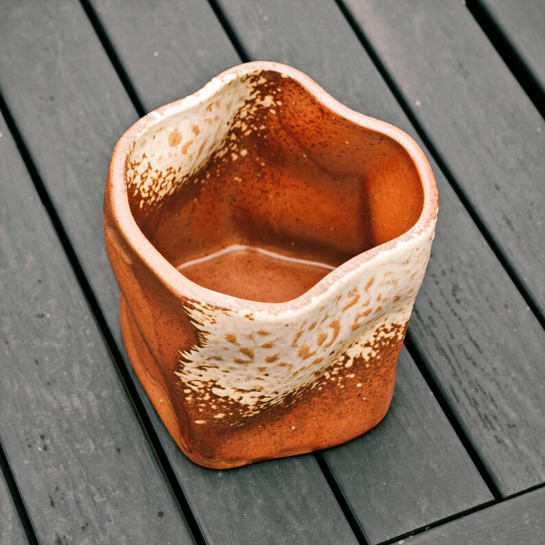 Peut inclure: Un vase en c&eacute;ramique fait &agrave; la main avec un design textur&eacute; unique. Le vase est d'une couleur orange-brun chaud et terreux avec un bord cr&egrave;me mouchet&eacute;. L'int&eacute;rieur est d'une nuance de brun plus fonc&eacute;e. Le vase a une forme irr&eacute;guli&egrave;re et l&eacute;g&egrave;rement froiss&eacute;e.