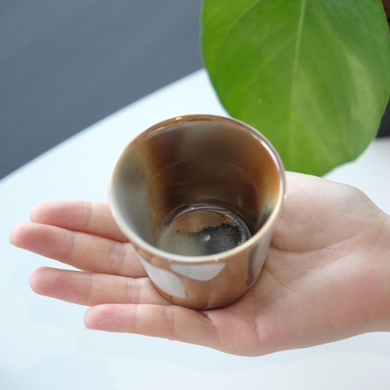 以下が含まれることがあります： 茶色と白の釉薬を施した、手作りの小さなセラミックカップ。カップは丸みを帯びた形で、人の手で持たれています。カップの内側は濃い茶色です。カップはおそらく飲用に使用されます。
